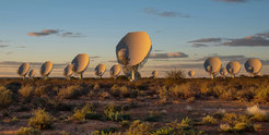 A row of large, white satellite dishes of the MeerKAT radio telescope in the South African semi-desert. The antennas stand in a dry, bushy landscape under a sky with soft clouds, while the evening sun creates a warm light atmosphere. The telescope is part of the Square Kilometre Array (SKA) and is used to explore the universe through radio astronomy.